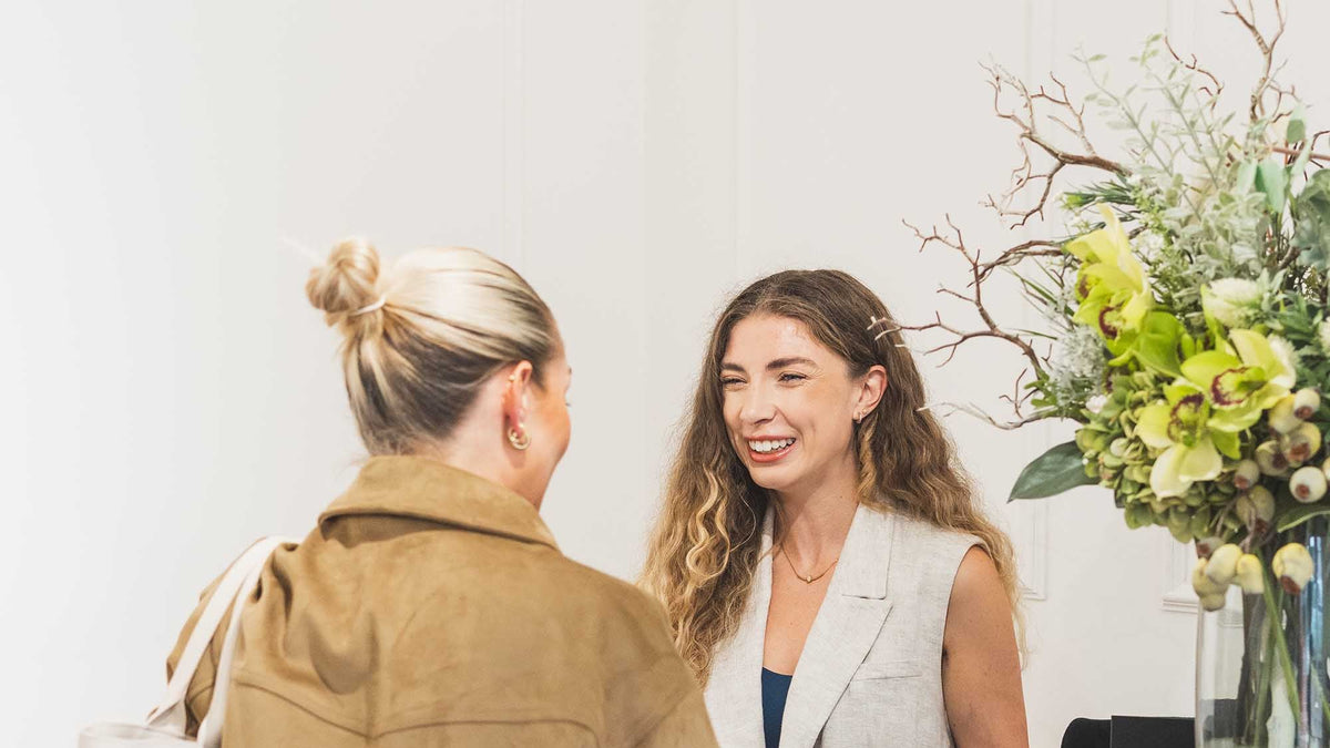Woman in a tan jacket greeting another woman at a reception desk with flowers in the background.