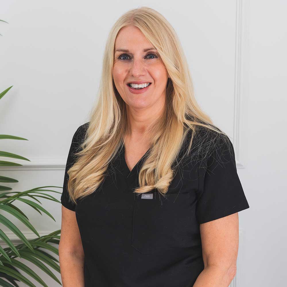 Woman with blonde hair wearing a black shirt in front of a white wall with a plant.