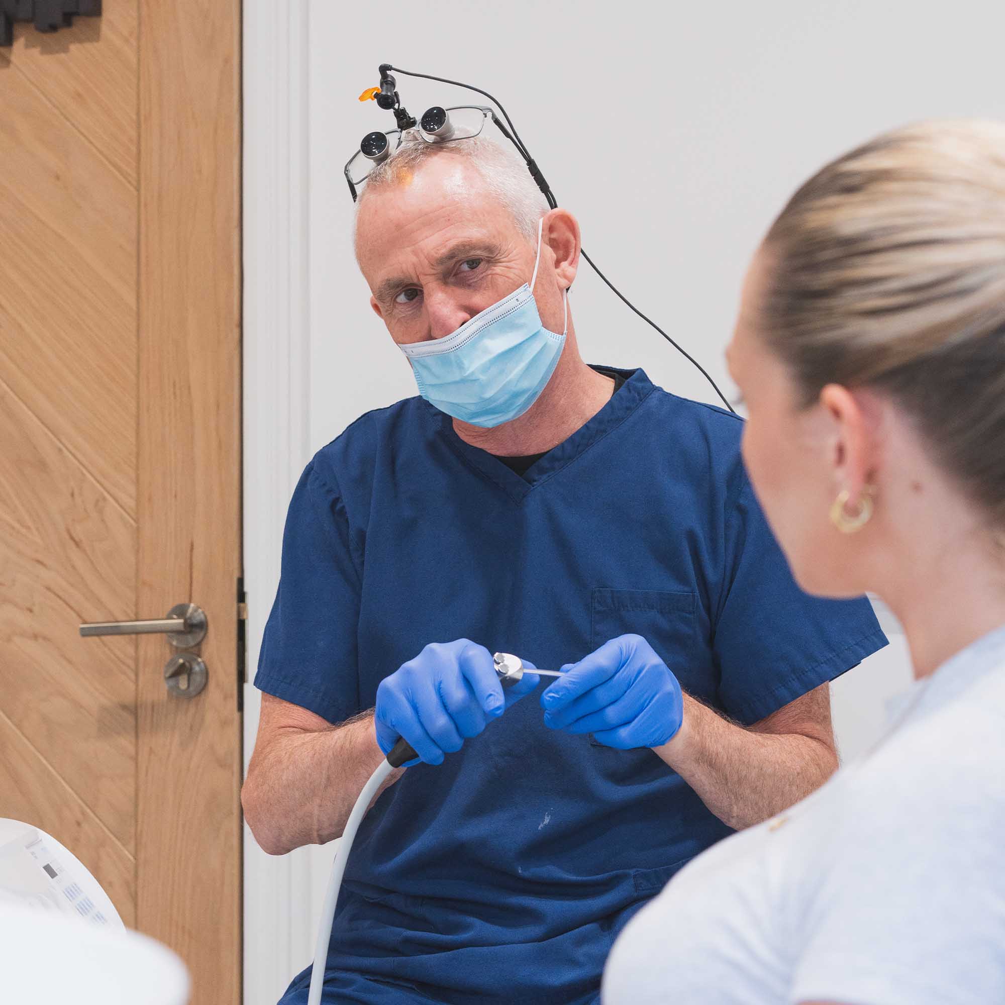 Dental professional in blue scrubs and gloves with a patient in a dental chair.