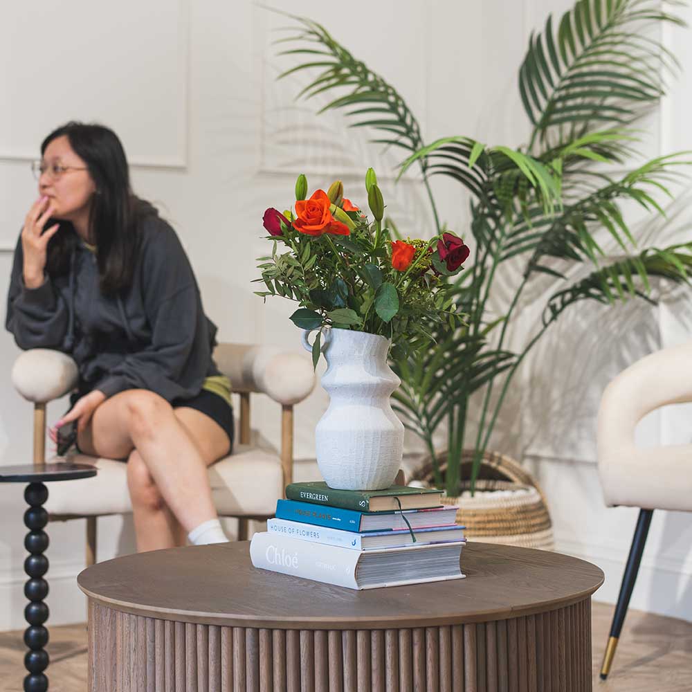 Woman sitting in a modern waiting room with a coffee table featuring books and flowers.
