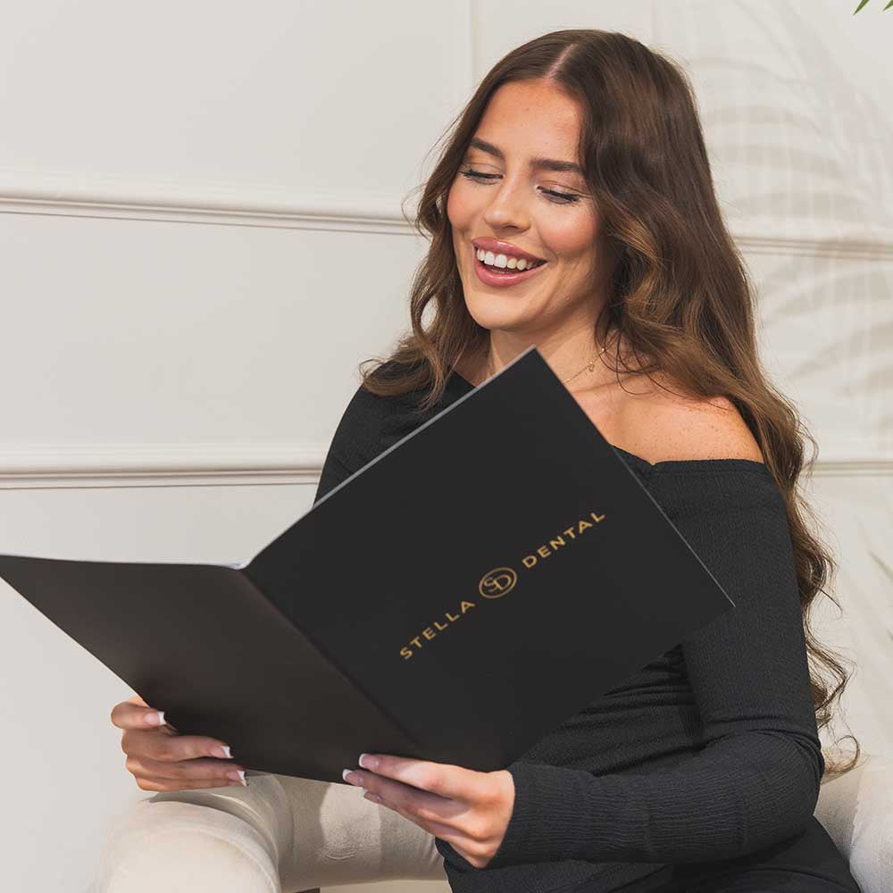 Woman holding a black folder with 'Stella Dental' logo against a white wall.